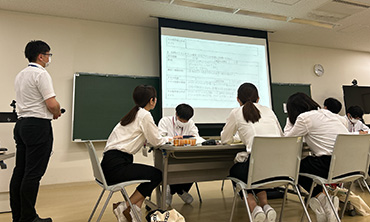 写真：長机に座っている生徒の横に立って、プロジェクターに写っている資料を説明している様子
